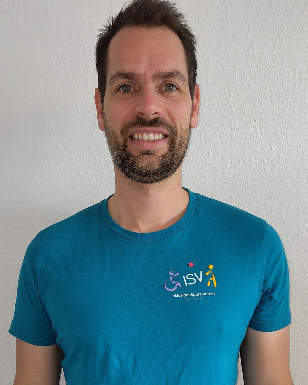 Ein Mann steht vor einer hellen Wand, lächelt freundlich und trägt ein ISV Alsterdorf T-Shirt.