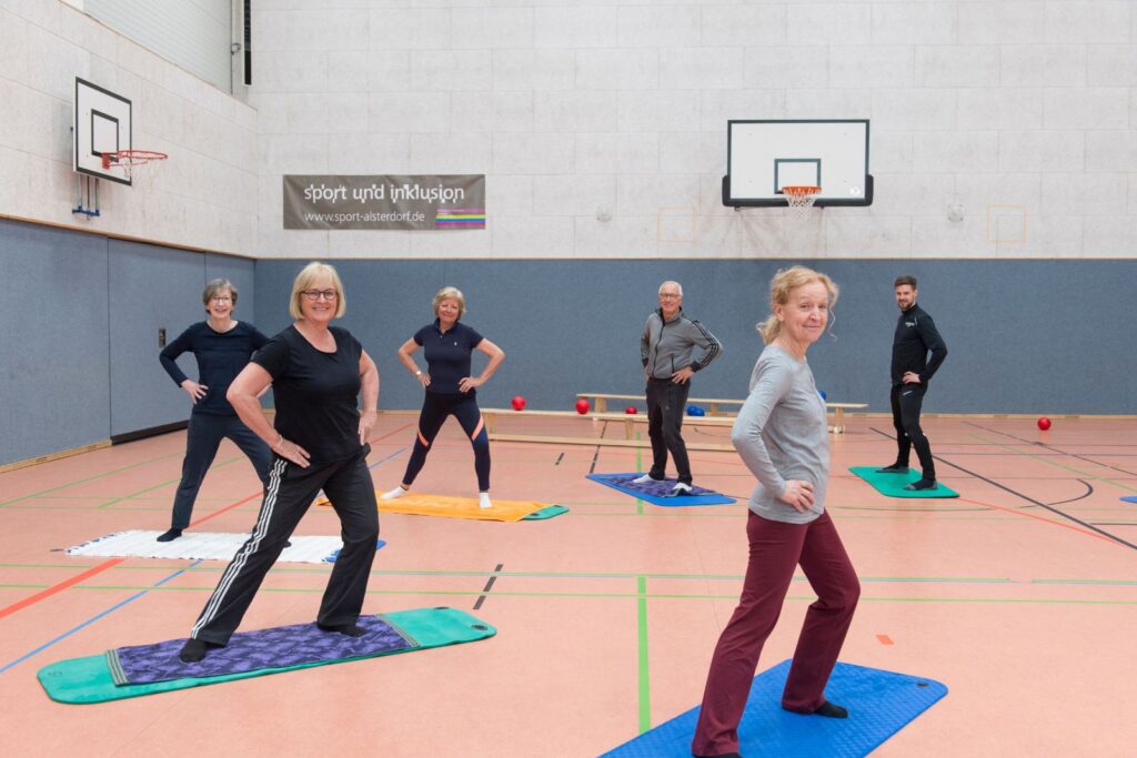 Eine Gruppe älterer Menschen übt gemeinsam Gymnastik auf Matten in der Sporthalle und lächelt in die Kamera.