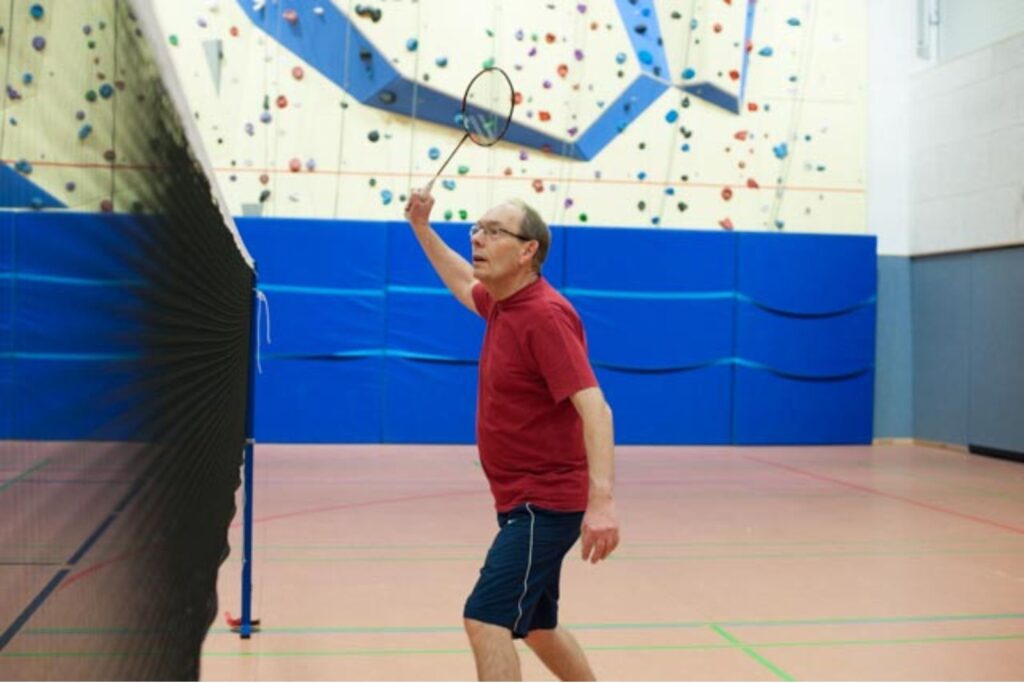 Ein Mann im roten Shirt spielt Badminton in der Halle und schlägt konzentriert den Ball über das Netz.
