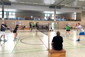 Mehrere Spielende spielen Badminton auf Feldern in einer großen Sporthalle.