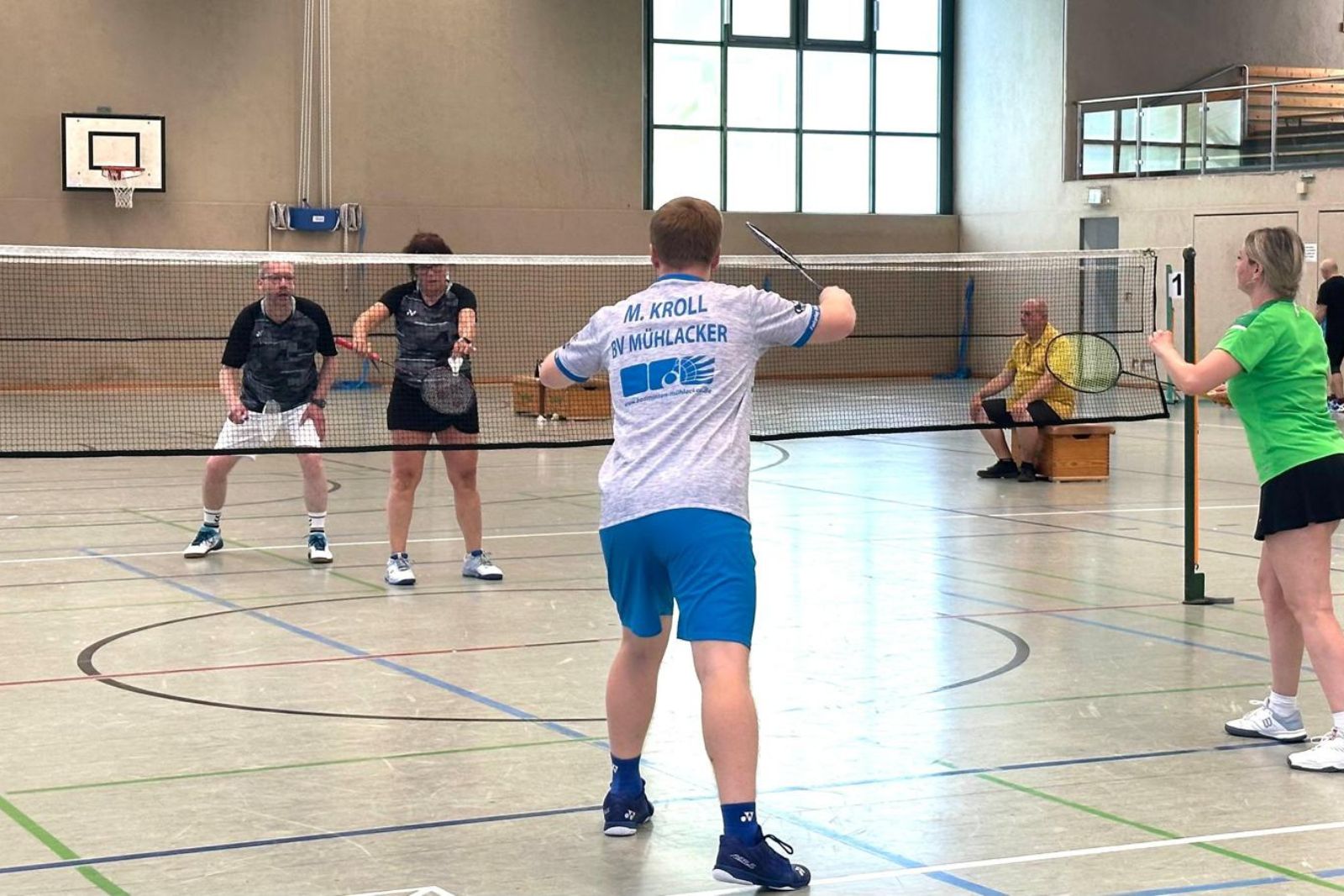 Zwei Teams spielen Badminton am Netz, eine Spielerin schlägt den Ball aus der Rückansicht.