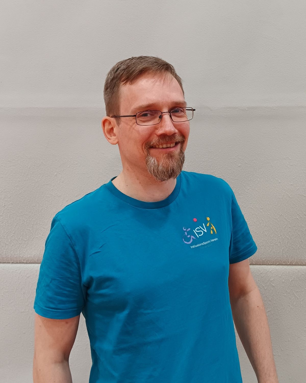 Ein Mann mit Brille und Bart lächelt in die Kamera und trägt ein türkisfarbenes ISV Alsterdorf Shirt.