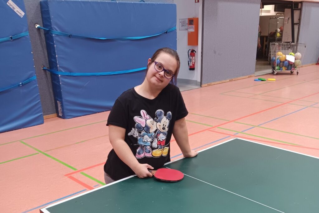 Person steht mit Tischtennisschläger an einer Tischtennisplatte in der Sporthalle.