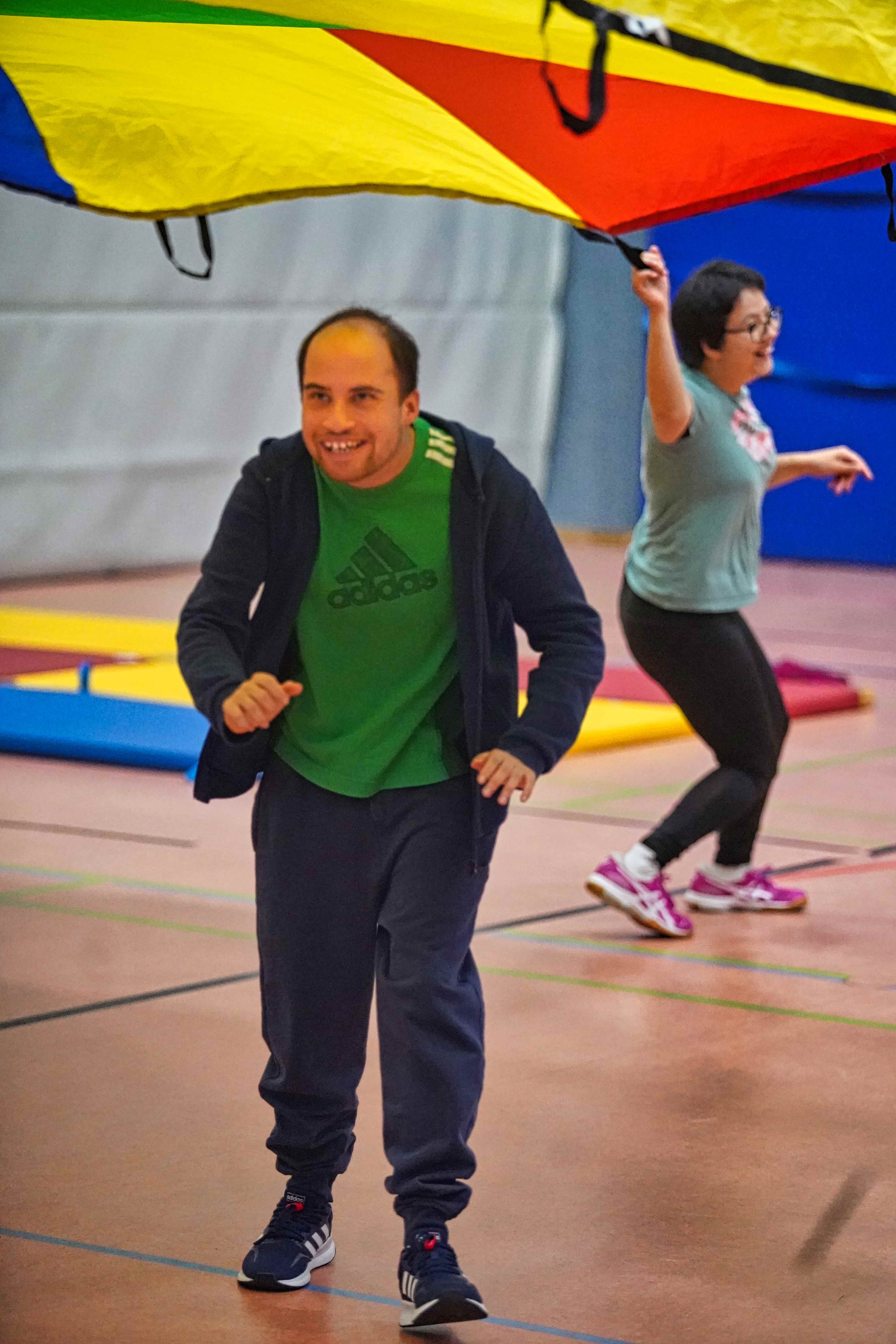 Ein Mann läuft lachend unter einem bunten Schwungtuch hindurch, während andere es in der Halle halten.