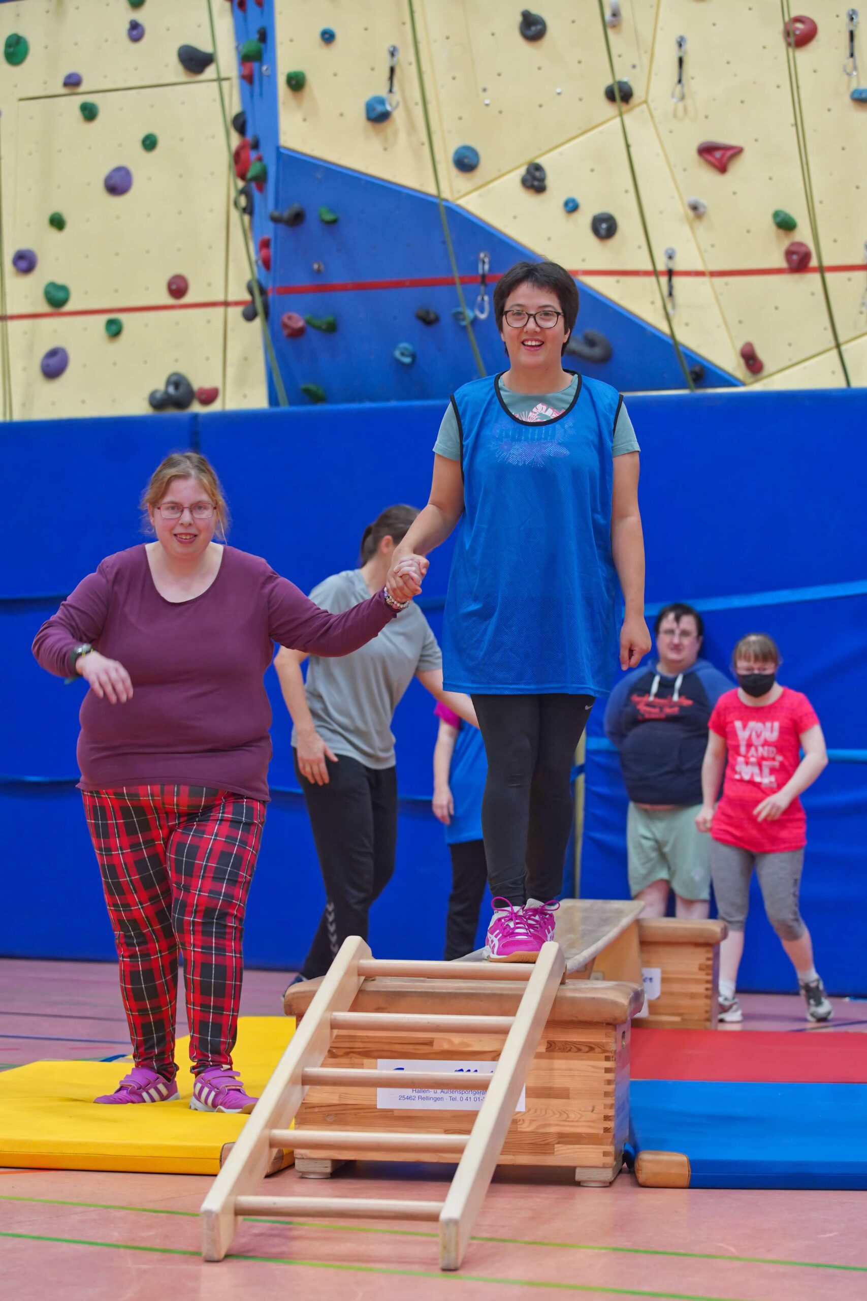 Zwei Frauen halten sich an den Händen, während eine über einen kleinen Holzsteg balanciert. Andere schauen zu.