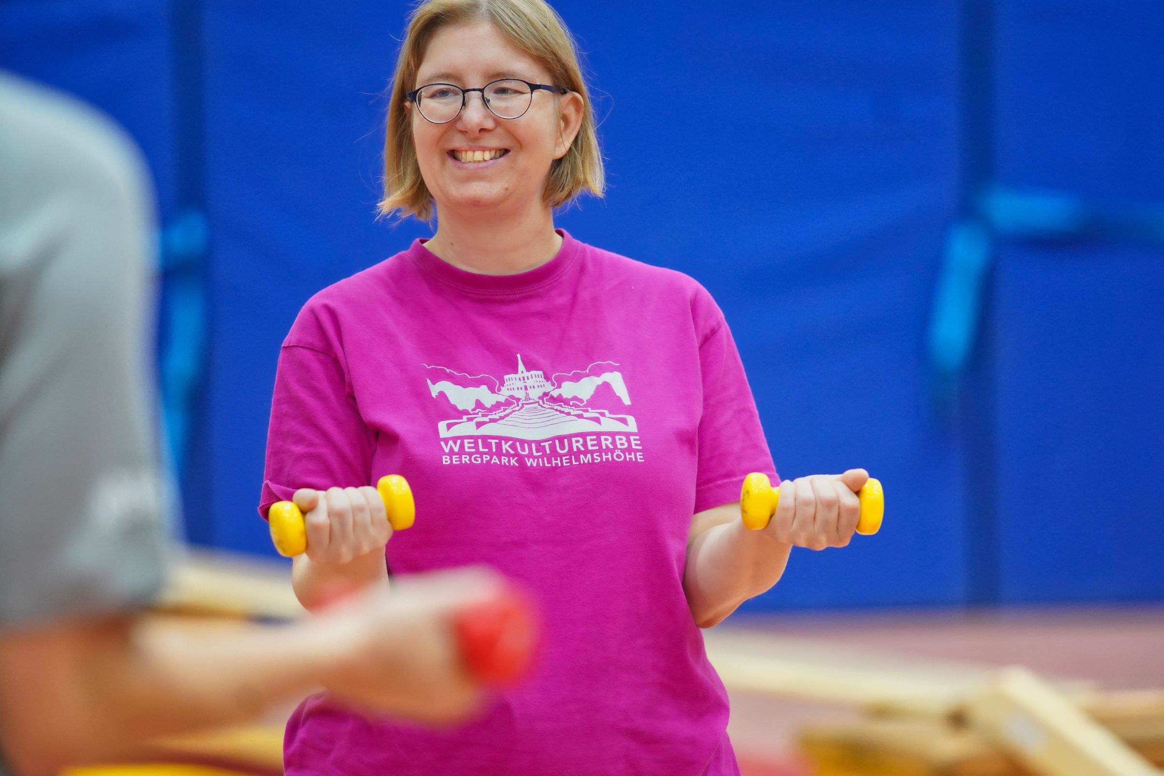 Eine Frau im pinken T-Shirt lächelt und hebt zwei gelbe Hanteln im gemeinsamen Training.