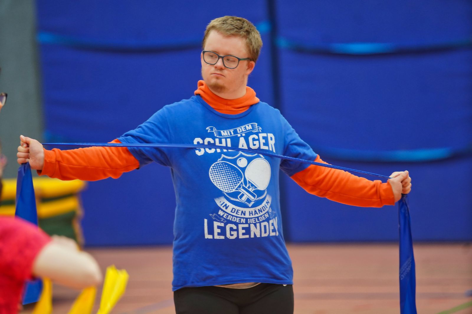 Ein junger Mann spannt ein blaues Widerstandsband zur Seite und übt konzentriert in der Sporthalle.