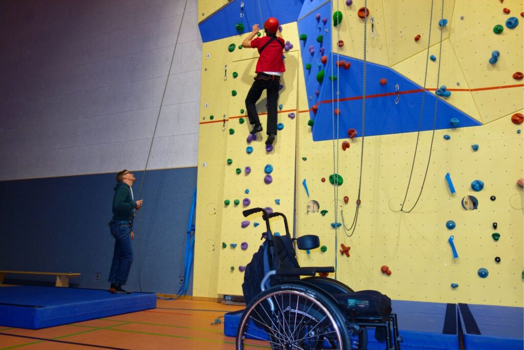 Eine Person klettert gesichert an der Kletterwand, während ein Trainer sichert. Ein Rollstuhl steht daneben.