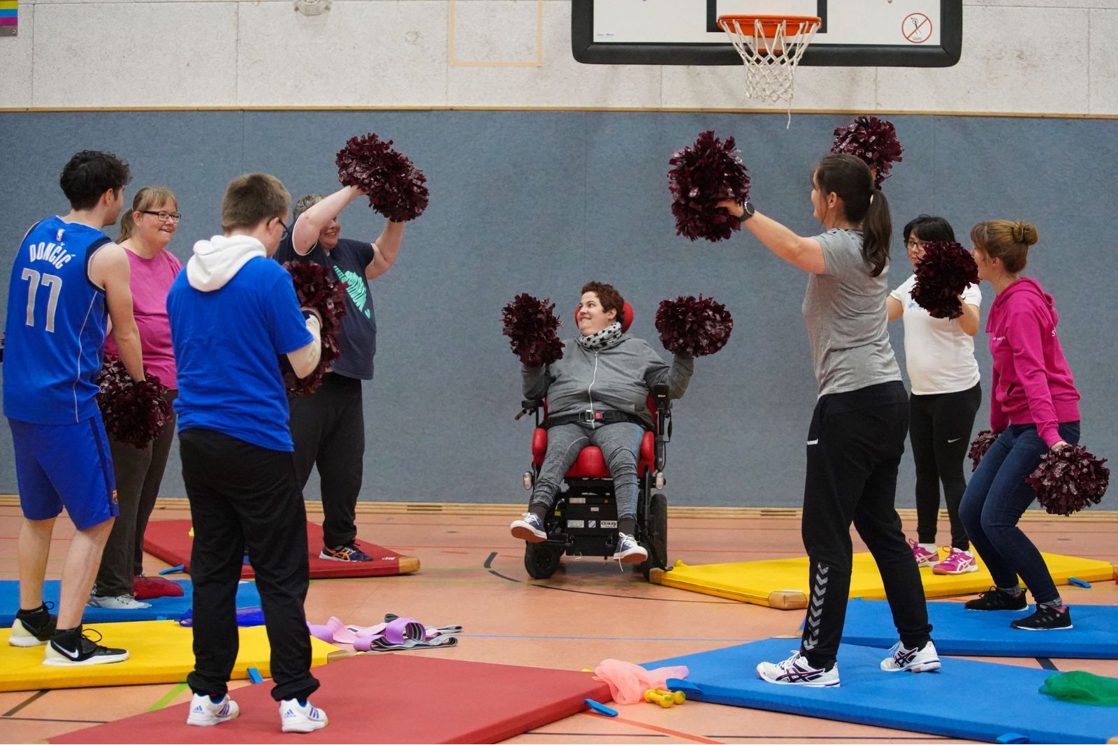 Eine Gruppe trainiert mit Pompons rund um eine Frau im Rollstuhl, die fröhlich am Training teilnimmt.