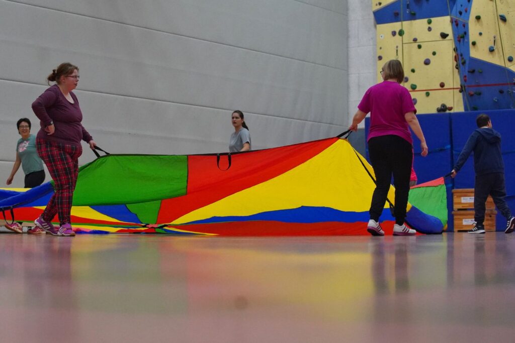 Mehrere Teilnehmende halten ein großes buntes Schwungtuch in der Sporthalle und bewegen es gemeinsam.