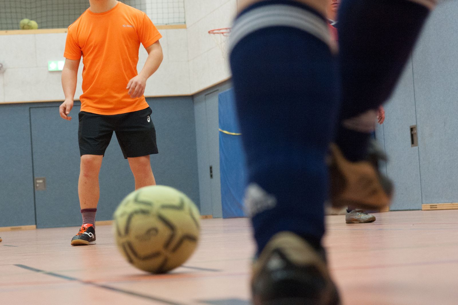 Fußball liegt im Vordergrund, mehrere Personen bewegen sich in einer Sporthalle während eines Trainings.