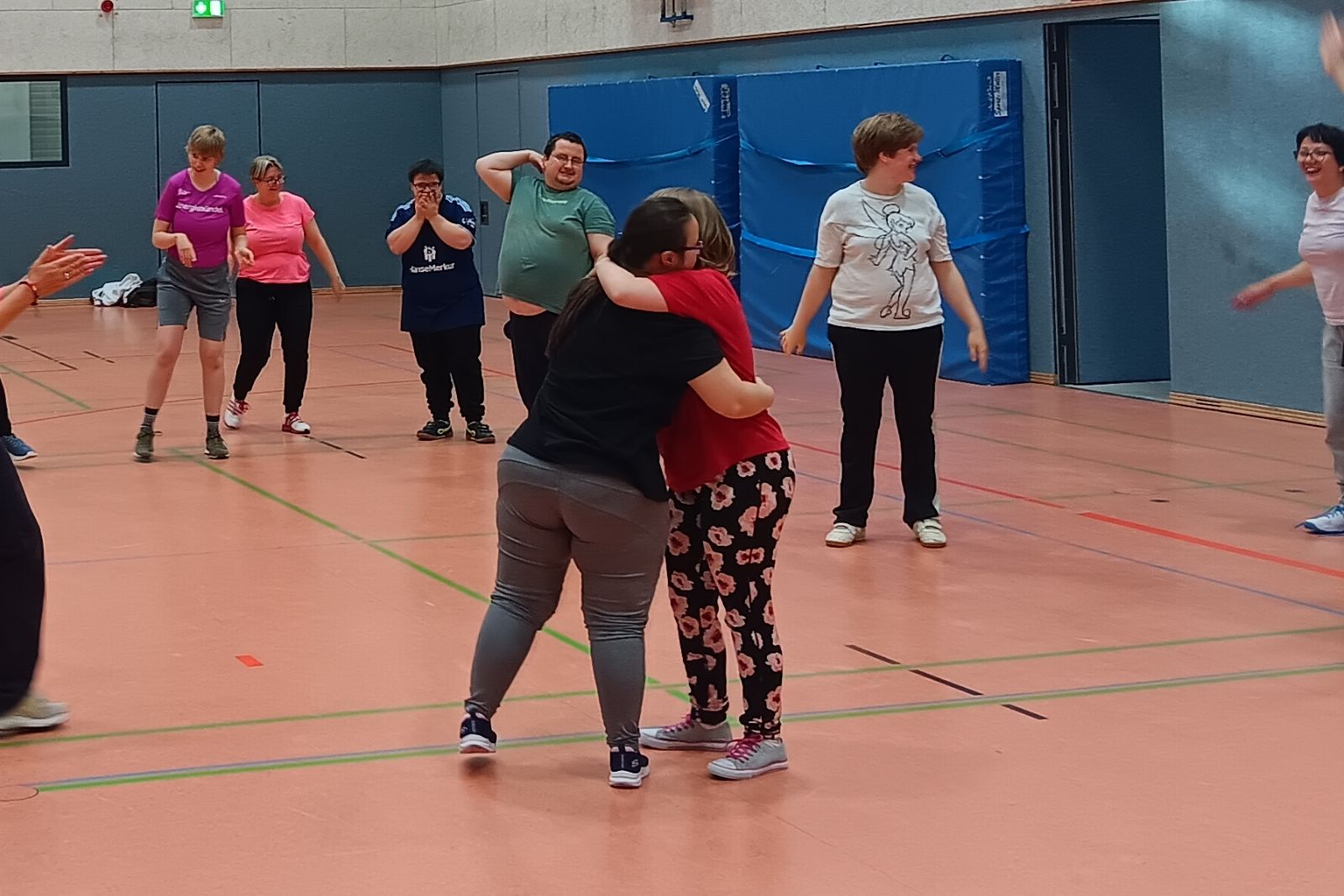 Zwei Personen umarmen sich in einer Sporthalle, andere Teilnehmende stehen gemeinsam im Hintergrund.