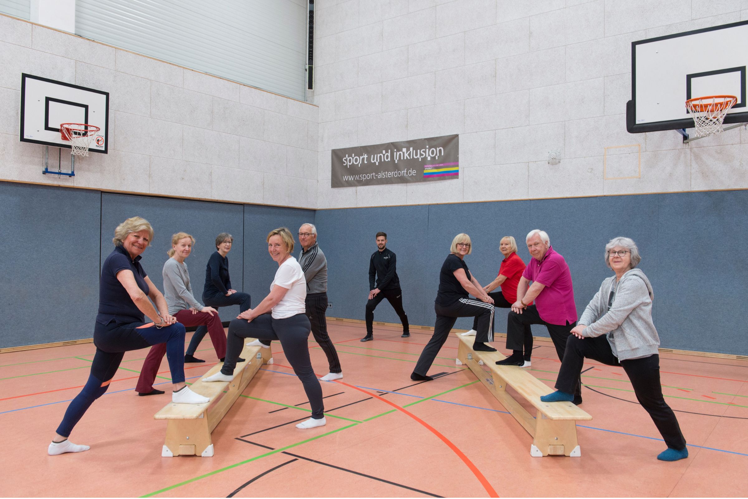 Mehrere ältere Erwachsene trainieren gemeinsam in einer Sporthalle auf Turnbänken, angeleitet von einem Trainer.