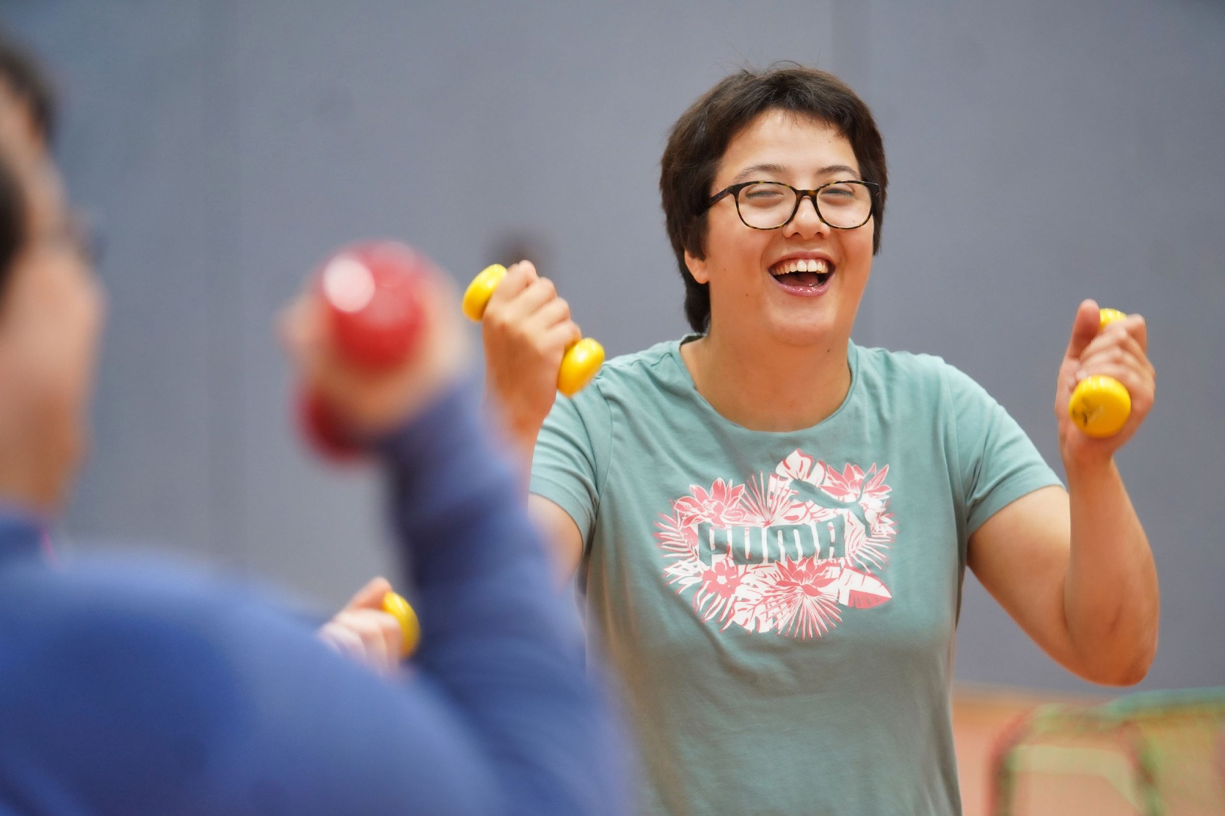 Eine Frau mit Brille lacht und trainiert mit gelben Hanteln gemeinsam in der Sporthalle.