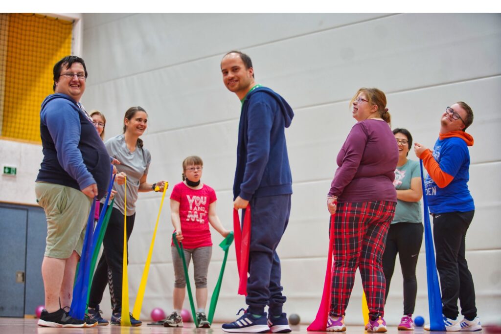 Eine gemischte Gruppe trainiert fröhlich mit bunten Widerstandsbändern in der Sporthalle.
