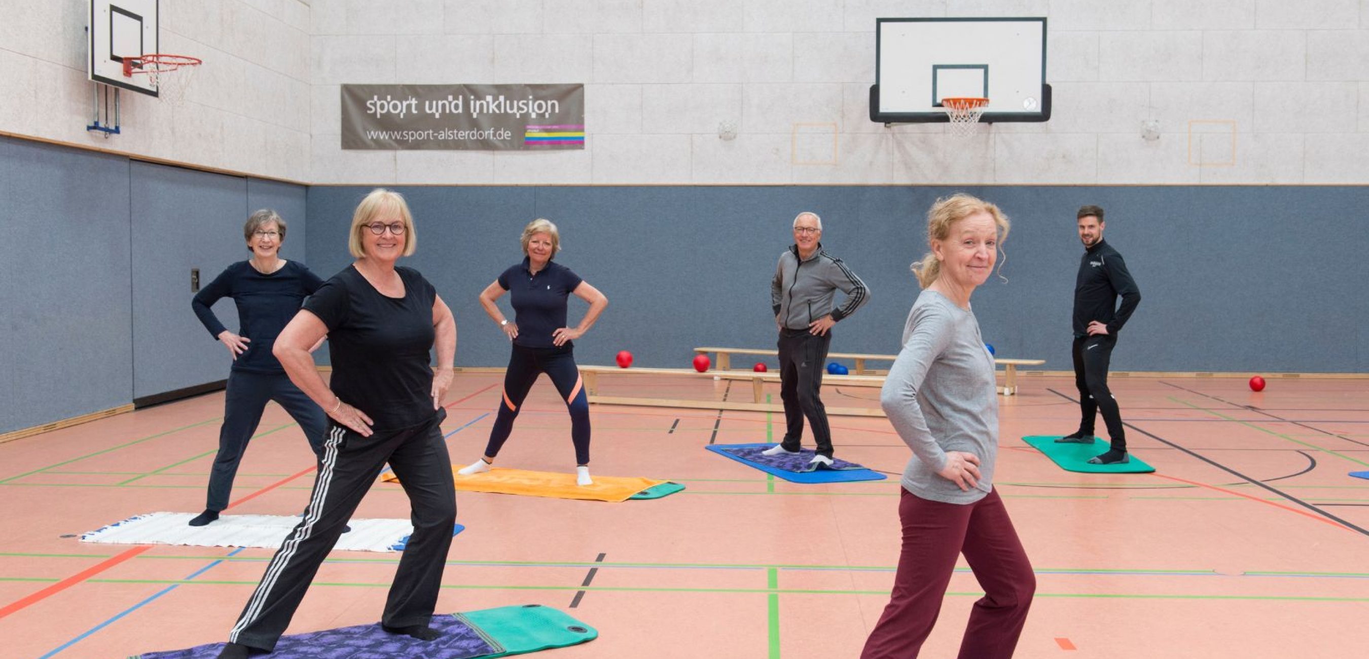 Eine Gruppe älterer Menschen übt gemeinsam Gymnastik auf Matten in der Sporthalle und lächelt in die Kamera.
