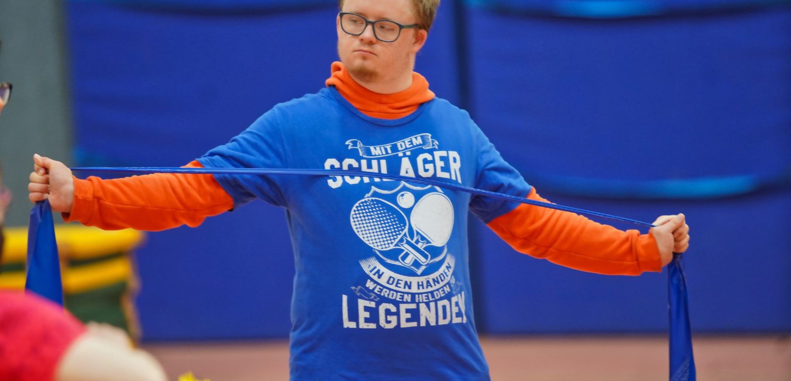 Ein junger Mann spannt ein blaues Widerstandsband zur Seite und übt konzentriert in der Sporthalle.