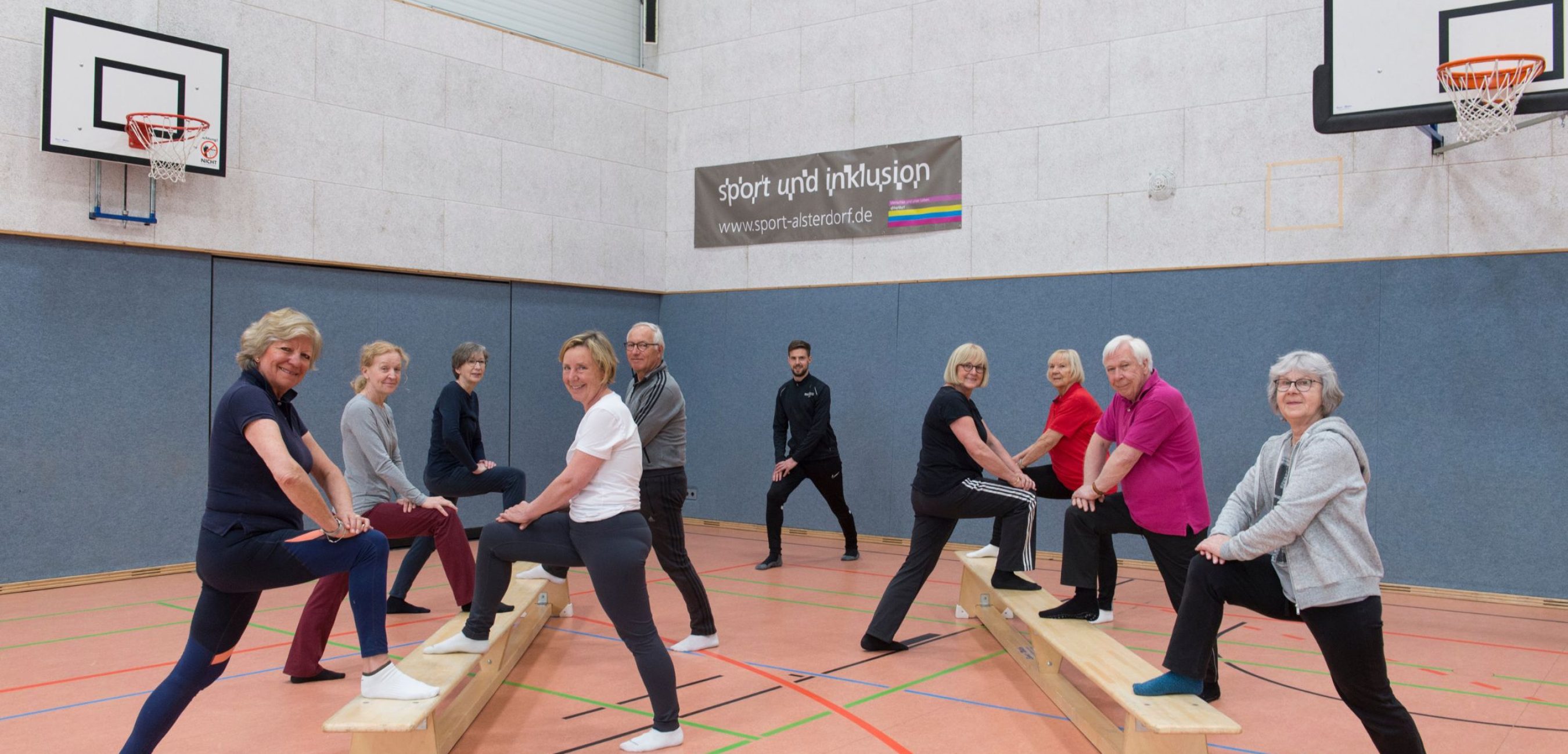 Mehrere ältere Erwachsene trainieren gemeinsam in einer Sporthalle auf Turnbänken, angeleitet von einem Trainer.