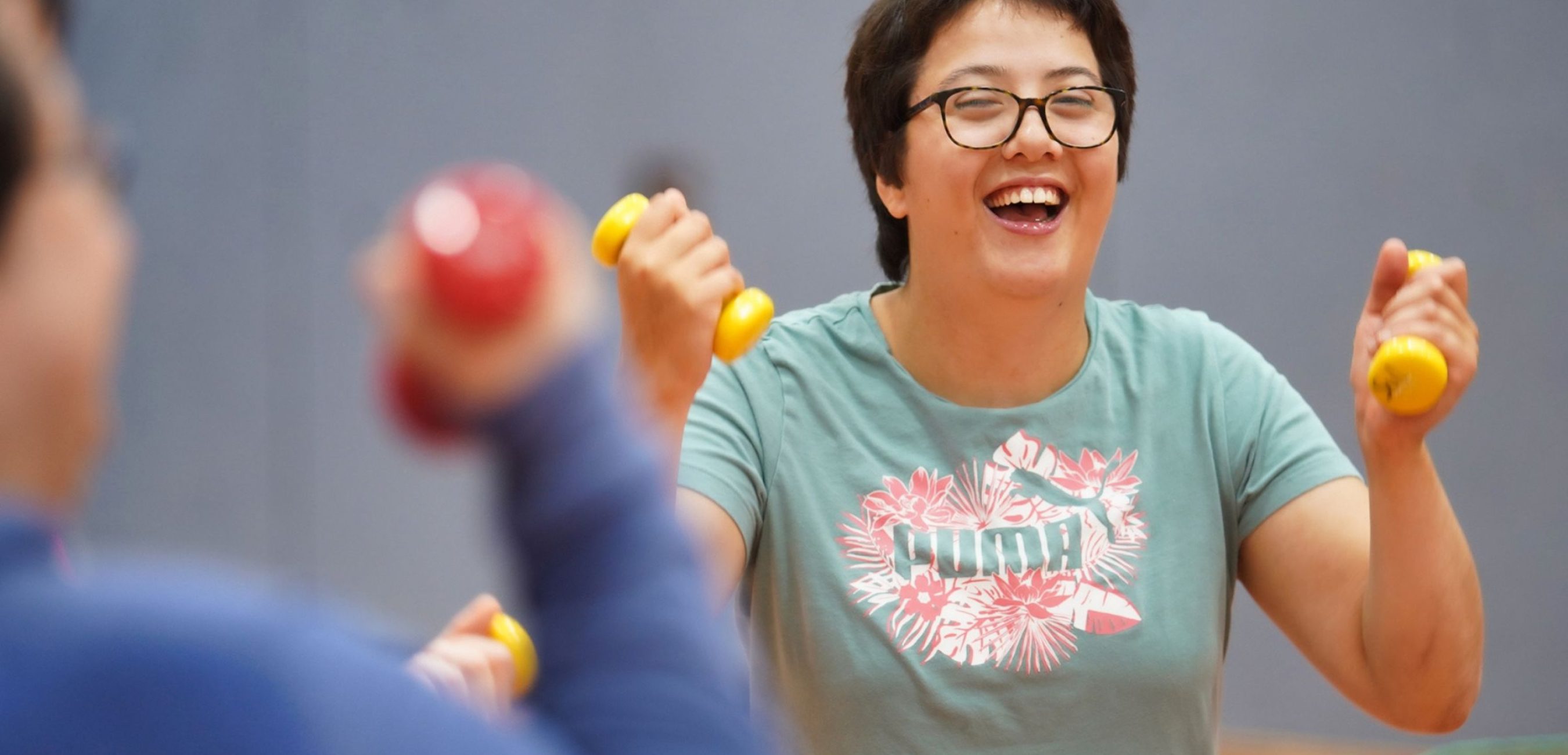 Eine Frau mit Brille lacht und trainiert mit gelben Hanteln gemeinsam in der Sporthalle.