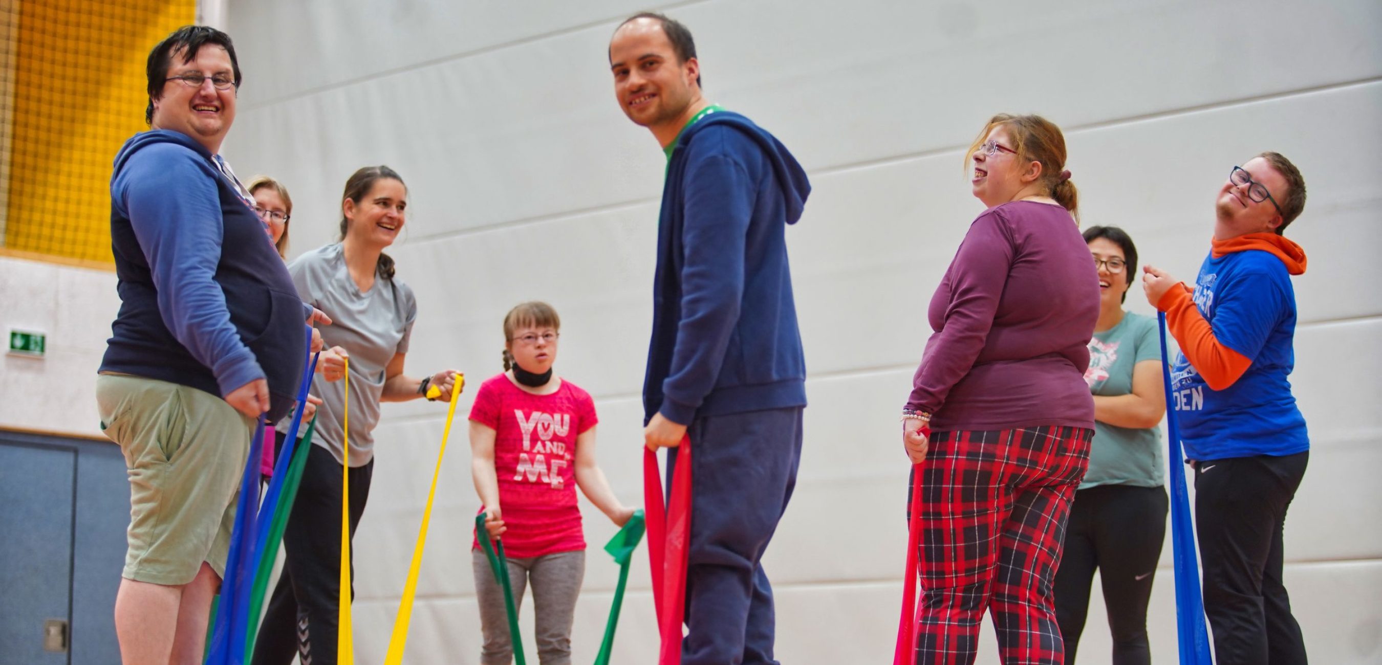 Eine gemischte Gruppe trainiert fröhlich mit bunten Widerstandsbändern in der Sporthalle.